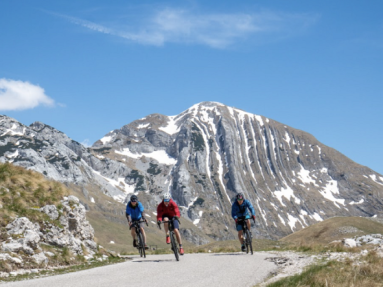 Drei Radfahrer auf einem Fahrradweg in den Bergen, im Hintergrund Bergpanorama. 