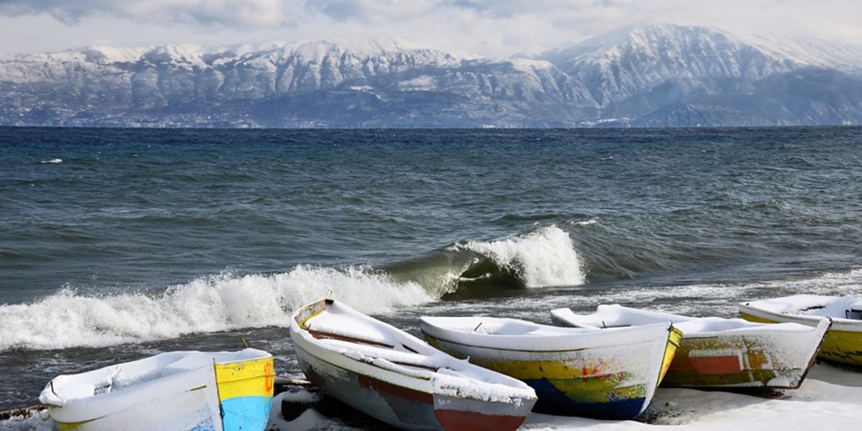 Im Vordergrund Fischerboote am Wasser, am hinteren Bildrand schneebedeckte Berge, dazwischen ein See