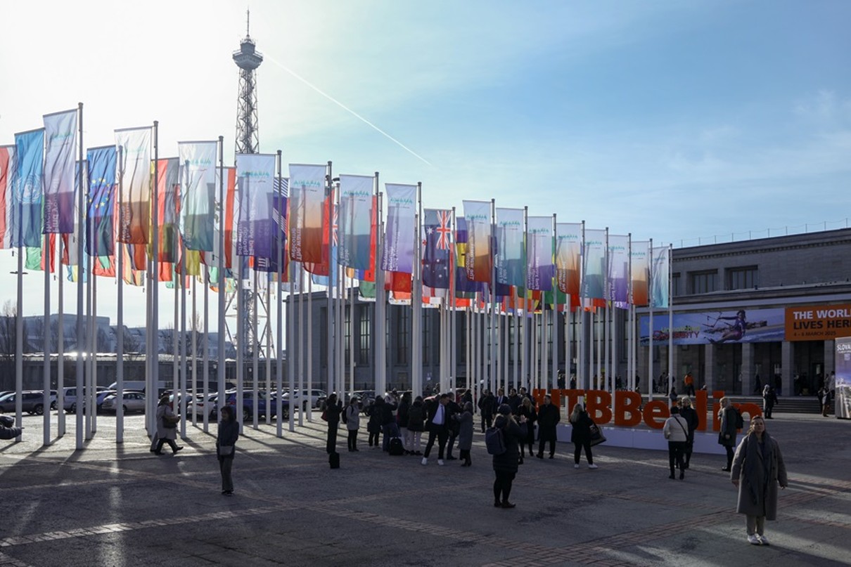 The image shows the outdoor area of the exhibition grounds, with numerous flags waving. In the background, the radio tower is visible.