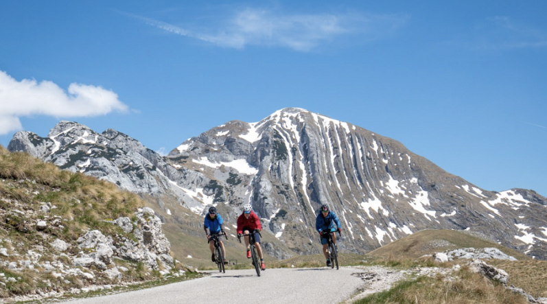 Drei Radfahrer auf einem Fahrradweg in den Bergen, im Hintergrund Bergpanorama. 