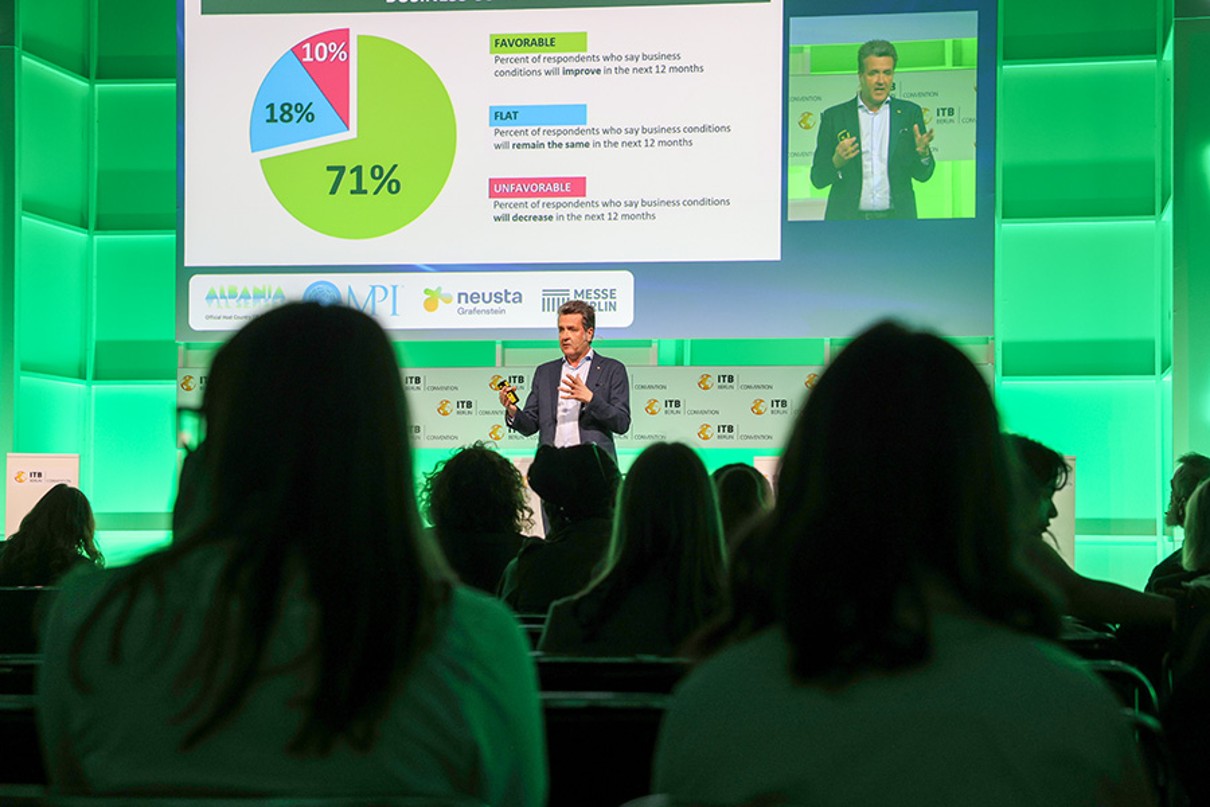 A man stands on stage and speaks to the audience, a presentation plays in the background