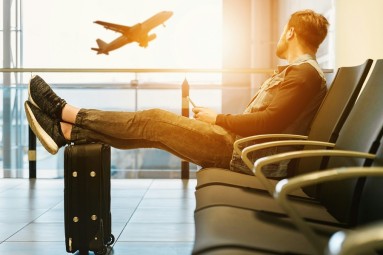A man sits in the check-in area of an airport; in the background an airplane takes off in the sunset.