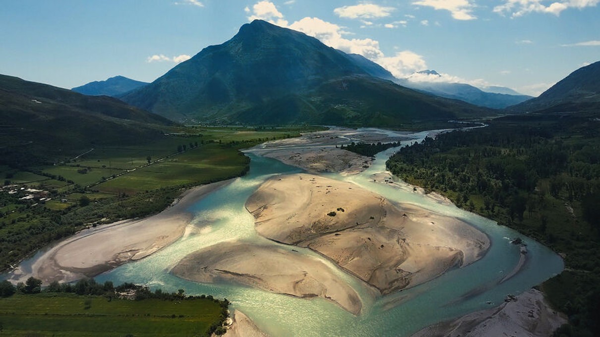 Flussdelta des Vjosa, mit einer Berglandschaft und grünen Feldern im Hintergrund.