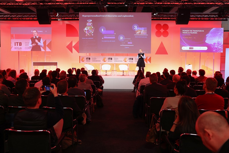 Audience in the foreground, directed towards a person on a stage holding a microphone. 